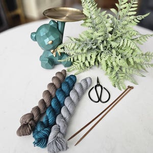 May include: A teal bear-shaped trinket holder with a gold-colored tray, next to a collection of yarn skeins in shades of brown, teal, and gray. Also included are a pair of scissors and knitting needles. A fern-like plant is in the background.