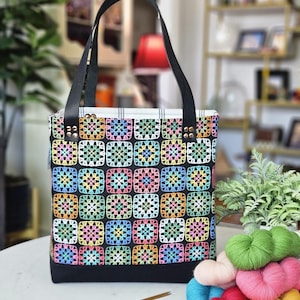 May include: A handmade tote bag with a colorful crochet pattern. The bag features a black base and leather handles. The crochet design incorporates squares of various colors, including pink, yellow, blue, and green. Knitting needles and yarn are also visible.