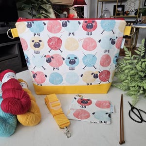 May include: A white knitting project bag with a sheep and yarn ball pattern, a yellow base, and a red zipper. A matching small pouch, yellow strap, and yarn are also visible. Knitting needles and scissors are also present.