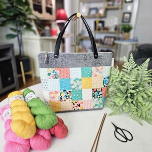 May include: A gray tote bag with black handles and a patchwork design on the front. The patchwork features squares of various colors and patterns. Beside the bag are colorful yarn skeins, knitting needles, and scissors.