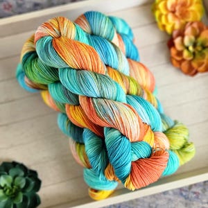 May include: A skein of colorful yarn in a braided pattern. The yarn features a blend of vibrant colors, including turquoise, orange, yellow, and green. The yarn is displayed on a white wooden tray, with decorative flowers and a succulent in the background.