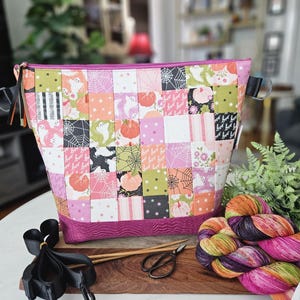 May include: A patchwork fabric project bag with a pink zipper and base, featuring Halloween-themed squares with ghosts, pumpkins, and spiderwebs. Knitting needles, scissors, and colorful yarn are on a wooden surface.