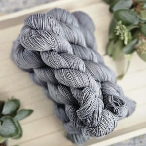 May include: A skein of yarn in a solid medium gray color with a twisted, braided texture. The yarn consists of multiple thin, tightly wound strands that create a consistent, smooth appearance throughout the entire length of the fiber bundle.