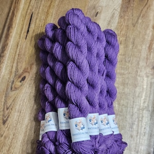 May include: A bundle of vibrant purple yarn skeins, neatly arranged on a wooden surface. Each skein is labeled with a small white tag featuring a logo. The yarn's color is a rich, saturated hue, perfect for knitting or crochet projects.