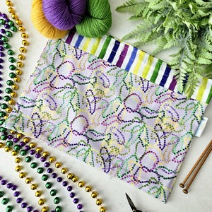 May include: A lilac fabric project bag with a dotted pattern in green, purple, and yellow. The bag is next to skeins of yellow, purple, and green yarn, Mardi Gras beads, scissors, and knitting needles.