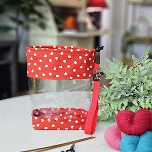 May include: A clear, rectangular knitting project bag with a red sheep pattern trim and a red wrist strap. Knitting needles, scissors, and colorful yarn balls are next to the bag. The bag is designed for storing knitting supplies.