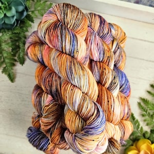 May include: A skein of hand-dyed yarn in a vibrant mix of colors. The yarn features a blend of orange, purple, and blue hues, creating a speckled effect. The yarn is arranged in a neat bundle, showcasing its texture and color variations. Ideal for knitting and crochet projects.