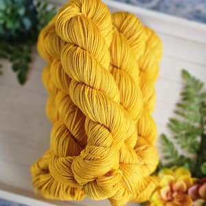 May include: A skein of bright yellow yarn, tightly wound and displayed on a white tray with greenery and a yellow flower. The yarn is a vibrant, solid color, perfect for knitting or crocheting projects.
