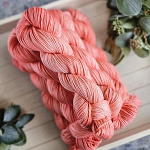 May include: A bundle of coral-colored yarn twisted into a skein. The yarn features a vibrant, solid pinkish-orange hue with a visible twisted fiber texture throughout the entire length of the material, showcasing a consistent and uniform color.