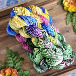 May include: A skein of hand-dyed yarn in a wooden tray. The yarn is a vibrant mix of colors, including yellow, blue, pink, purple, and green. The yarn is arranged in a twisted bundle. Decorative flowers and greenery are in the background.