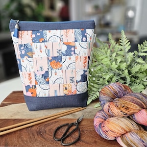 May include: A patchwork fabric yarn bag with a blue denim base and trim, featuring a floral and dreamcatcher pattern. Knitting needles, scissors, and a skein of colorful yarn are next to the bag. An owl figurine and greenery are in the background.