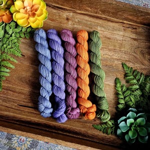 May include: A wooden tray displays six skeins of colorful yarn in shades of blue, purple, orange, and green. The yarn is arranged in a row, with decorative flowers and greenery on the sides. The yarn is likely for knitting or crochet projects.