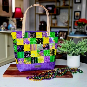May include: A purple and green patchwork tote bag with leather handles. The bag is decorated with a Mardi Gras theme and has a zipper closure. The bag is sitting on a wooden surface with a string of Mardi Gras beads.