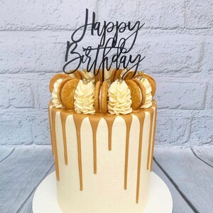 Op de afbeelding: Een witte cake met een bruine drip-glazuur en een "Happy Birthday"-topper. De cake is versierd met gouden sprinkles en ronde koekjes.