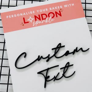Könnte beinhalten: Eine weiße Karte mit den Worten "Custom Text" in schwarzer Schreibschrift. Die Karte hat eine rosa und weiße Kopfzeile mit dem Text "Personalise Your Bakes With London Sparkle". Die Karte liegt auf einem schwarzen Drahtgestell.