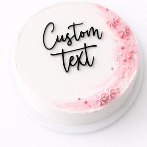 Puede incluir: Una tarta redonda blanca con acentos de glaseado rosa y decoraciones florales. La tarta presenta las palabras "Custom text" en escritura negra. La tarta está sobre un plato blanco.