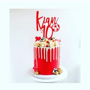 Könnte beinhalten: Roter und weißer Kuchen mit einem roten und weißen Kuchentopper, auf dem "Kian 10" mit einem Fußball steht. Der Kuchen ist mit weißem Frosting und roten und weißen Streuseln verziert.