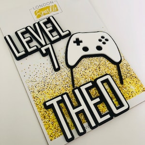May include: White cake topper with black outlines. The topper says "Level 7 Theo" and includes a white video game controller with black buttons.