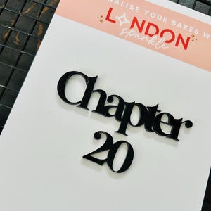 Könnte beinhalten: Schwarzer Kuchenaufsatz aus Acryl mit der Aufschrift "Chapter 20". Der Aufsatz befindet sich auf einem weißen Hintergrund mit einem Banner mit der Aufschrift "Personalise your bakes with London Sparkle".