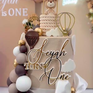 May include: A decorated cake with the text "Leyah Turns One". The cake features a teddy bear in a hot air balloon, gold and white balloons, and decorative elements. The cake is beige with white and gold accents.