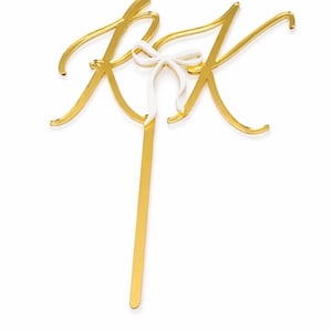 Peut inclure: Un cake topper doré avec les lettres entrelacées "KK" et un délicat nœud blanc. Le cake topper a une longue tige fine pour être inséré dans un gâteau. Ce design élégant convient aux mariages ou autres célébrations.