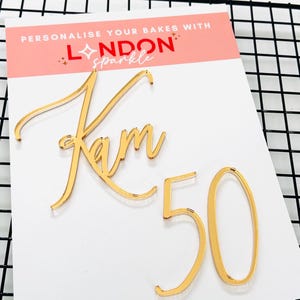 May include: A gold cake topper featuring the name "Kam" and the number "50". The topper is made of a shiny material and is designed for cakes. The text "PERSONALISE YOUR BAKES WITH LONDON sparkle" is printed on a pink background.