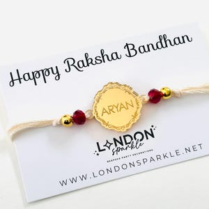 May include: A gold bracelet with the name "ARYAN" on a decorative charm. The bracelet has red and gold beads and is presented on a card that reads "Happy Raksha Bandhan". The card also displays the "LONDON sparkle" logo.