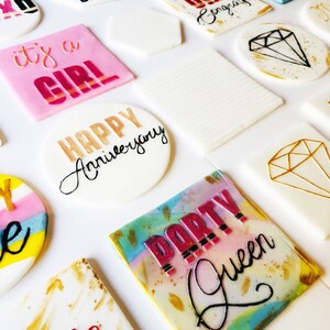 Puede incluir: Surtido de adornos para pasteles redondos y cuadrados con varios colores y dise&ntilde;os. Los adornos incluyen texto como "Live Laugh Love", "Hello Good Times", "It's a Girl", "Happy Anniversary" y "Party Queen".