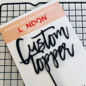 Op de afbeelding: Zwarte 'Custom Topper' taartdecoratie met een lange steel. De topper staat op een witte kaart met de tekst 'Personalise your bakes with London Sparkle' in rood en zwart. De achtergrond is een zwart draadrek.