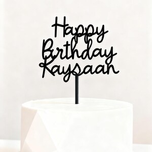 Op de afbeelding: Een zwarte taarttopper met de woorden "Happy Birthday Kaysaan" in een cursief lettertype. De topper is geplaatst op een witte taart met een geometrisch ontwerp.