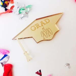 May include: Gold graduation cap shaped acrylic ornament with the text "GRAD of 2020" in a gold script font.