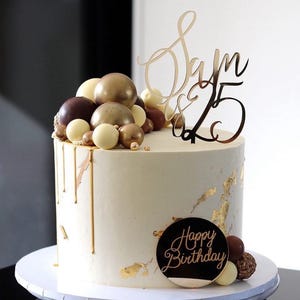 May include: A white cake with gold drip icing and gold leaf accents. The cake is decorated with chocolate and white chocolate balls and a gold mirror acrylic topper that says "Sam & 25" and "Happy Birthday".