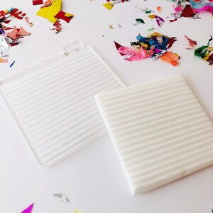 Puede incluir: Dos superficies cuadradas de textura blanca con l&iacute;neas en relieve sobre un fondo blanco. Las superficies est&aacute;n rodeadas de confeti de colores.