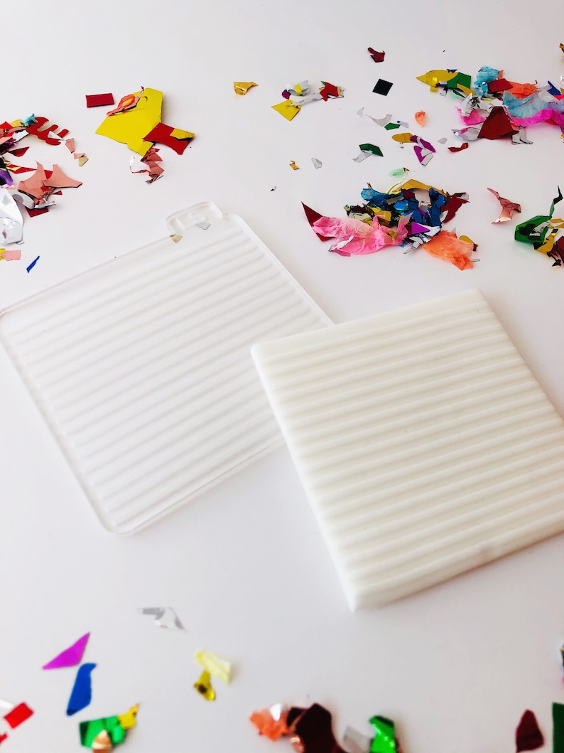 Puede incluir: Dos superficies cuadradas de textura blanca con l&iacute;neas en relieve sobre un fondo blanco. Las superficies est&aacute;n rodeadas de confeti de colores.