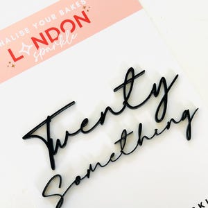 Puede incluir: Adorno negro para tarta "Twenty Something" en fuente cursiva. El adorno está sobre una superficie blanca con una tarjeta rosa y blanca en el fondo que dice "London Sparkle".