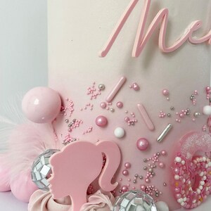 Pode incluir: Um bolo degrad&ecirc; rosa e branco decorado com confeitos rosa e prateados, bolas rosa e brancas, uma silhueta de Barbie rosa e duas bolas de discoteca prateadas. O bolo &eacute; coroado com uma inscri&ccedil;&atilde;o rosa em cursiva que diz "Mia".
