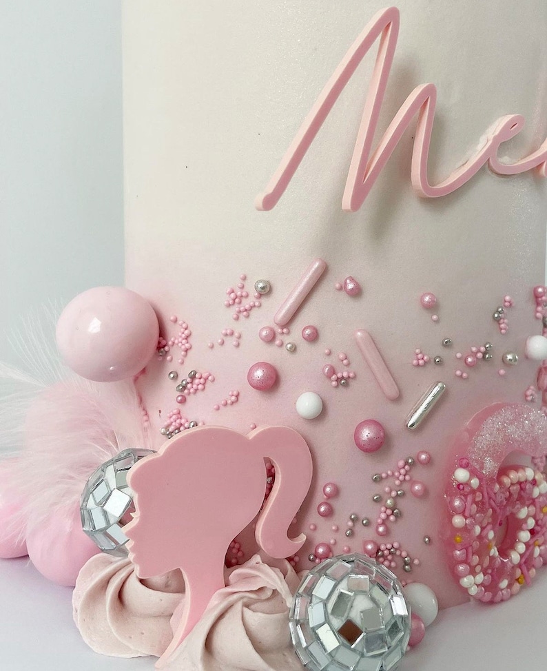 Pode incluir: Um bolo degrad&ecirc; rosa e branco decorado com confeitos rosa e prateados, bolas rosa e brancas, uma silhueta de Barbie rosa e duas bolas de discoteca prateadas. O bolo &eacute; coroado com uma inscri&ccedil;&atilde;o rosa em cursiva que diz "Mia".