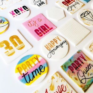 Puede incluir: Surtido de adornos para pasteles redondos y cuadrados de colores con diferentes frases y dise&ntilde;os, incluyendo "Hello good times", "Live laugh love", "Party time", "It's a girl", "Happy Anniversary", "Congrats", "Party Queen" y "Love".