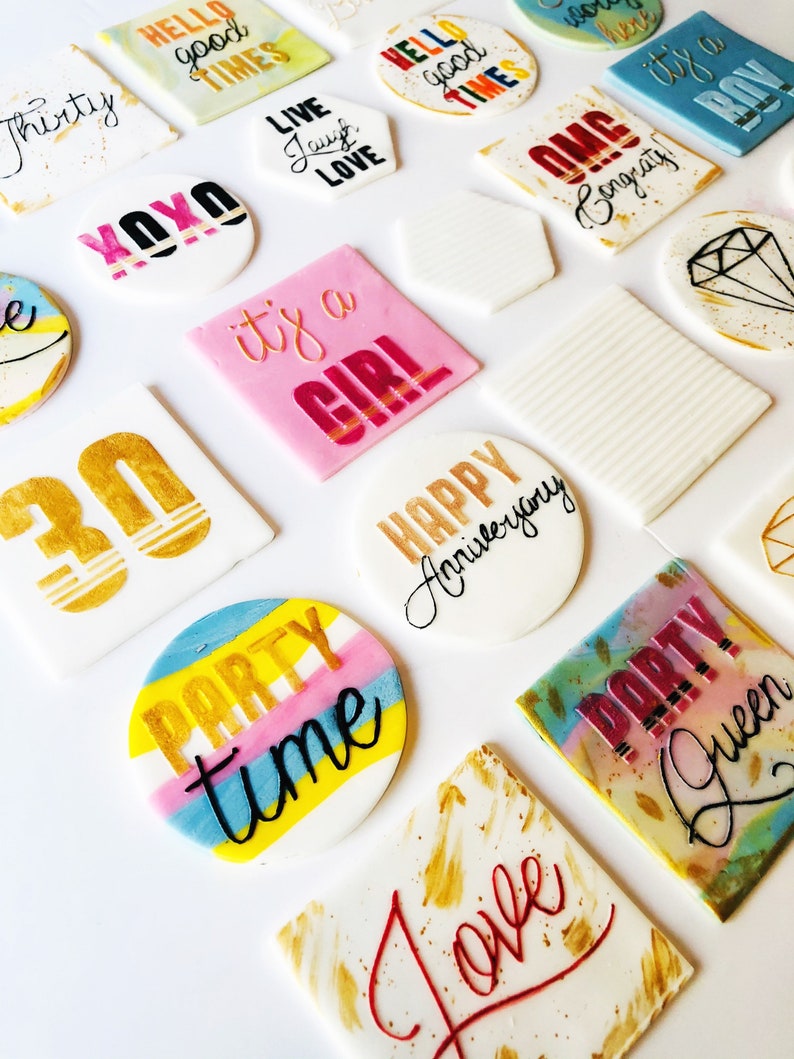 Puede incluir: Surtido de adornos para pasteles redondos y cuadrados de colores con diferentes frases y dise&ntilde;os, incluyendo "Hello good times", "Live laugh love", "Party time", "It's a girl", "Happy Anniversary", "Congrats", "Party Queen" y "Love".