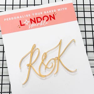 Könnte beinhalten: Ein goldfarbener Tortenaufsatz mit den Buchstaben "R&K" in Schreibschrift. Der Aufsatz befindet sich auf einer weißen Karte mit dem Text "PERSONALIZE YOUR BAKES WITH LONDON sparkle" in Rot und Weiß.