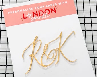 May include: A gold-colored cake topper with the letters "R&K" in a cursive font. The topper is on a white card with the text "PERSONALIZE YOUR BAKES WITH LONDON sparkle" in red and white.