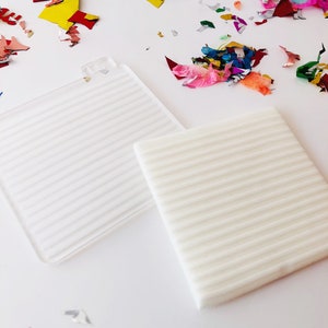 Puede incluir: Dos posavasos cuadrados blancos texturados con un patr&oacute;n acanalado. Los posavasos est&aacute;n sobre una superficie blanca con confeti de colores esparcidos a su alrededor.