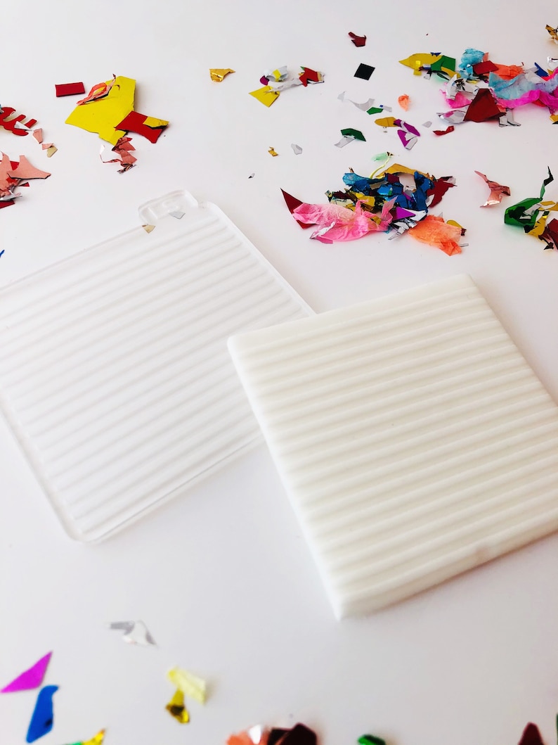 Puede incluir: Dos posavasos cuadrados blancos texturados con un patr&oacute;n acanalado. Los posavasos est&aacute;n sobre una superficie blanca con confeti de colores esparcidos a su alrededor.