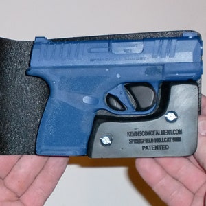 May include: A blue handgun with a black holster. The handgun is held in place by a gray base with the text "KEVINSCONCEALMENT.COM SPRINGFIELD HELLCAT 9MM PATENTED". The holster is black and textured.