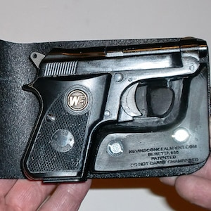 May include: A black Beretta 950 pistol is partially inserted into a black leather wallet. The pistol has a silver trigger and a circular emblem with the letters "WE". The wallet has text that reads "KEVINSCONCEALMENT.COM BERETTA 950 PATENTED DO NOT CARRY CHAMBERED".