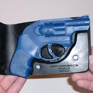 May include: A black leather wallet with a blue handgun holster. The blue handgun is partially visible, with the text "RUGER LCR" and "KEVINSCONCEALMENT.COM" on the holster. The wallet is held open, showcasing the holster's design.