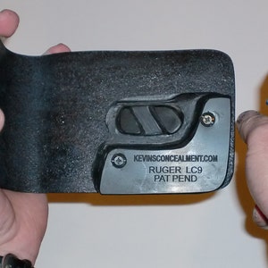 May include: A black leather holster for a Ruger LC9 pistol. The holster has a metal plate with the text "KEVINSCONCEALMENT.COM RUGER LC9 PAT PEND".