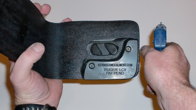 May include: A black leather holster for a Ruger LC9 pistol. The holster has a metal plate with the text "KEVINSCONCEALMENT.COM RUGER LC9 PAT PEND".