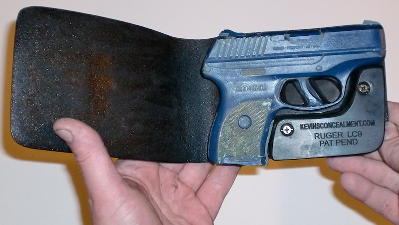 May include: A blue Ruger LC9 pistol is held in a black leather holster. The holster has a metal plate with the text "KEVINSCONCEALMENT.COM RUGER LC9 PAT PEND".