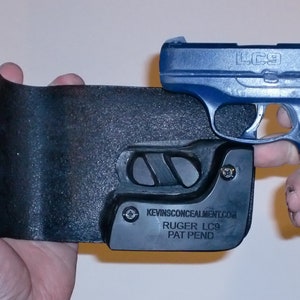 May include: A black leather holster with a blue Ruger LC9 pistol inside. The holster has the text "KEVINSCONCEALMENT.COM" and "RUGER LC9 PAT PEND" on it.